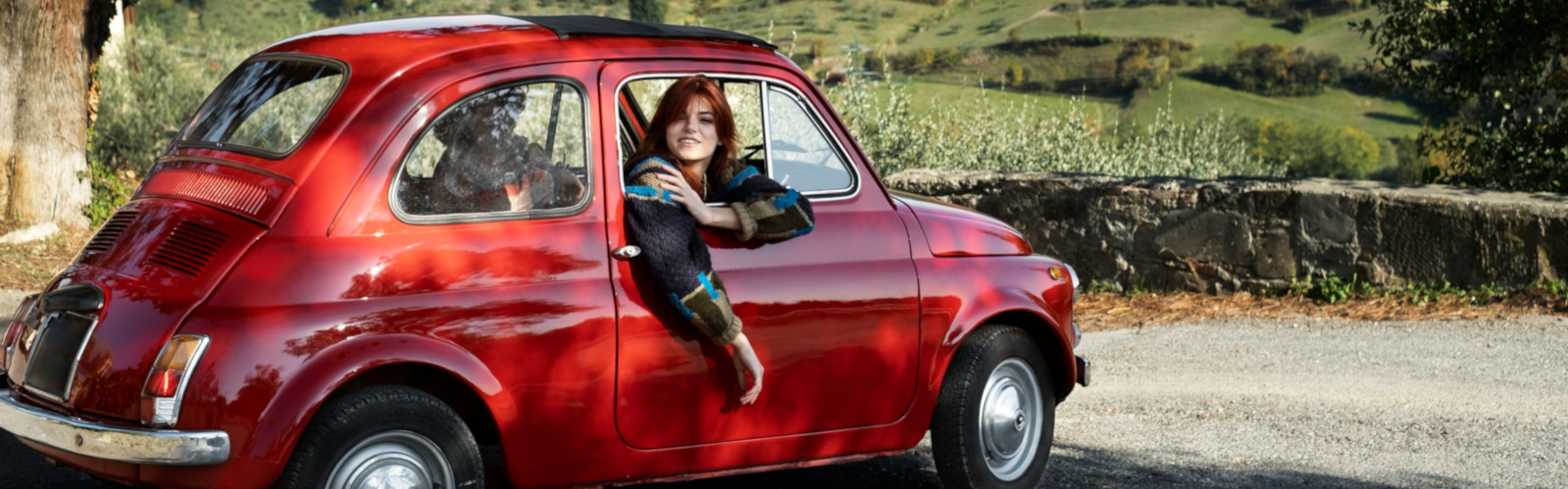 A woman driving a red vintage Fiat through the Italian countryside, surrounded by rolling green hills and olive groves.